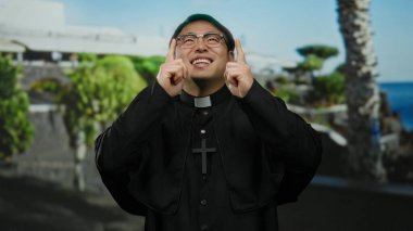 Papaz kılığındaki Asyalı adam gülümsüyor ve arka planda yeşillik ve okyanusla güneşli bir sahil geçidinde yukarı doğru hareket ediyor..