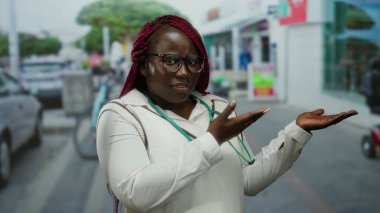 Örgülü bir kadın, gözlüklü ve bluzlu bir şekilde gülümsüyor şehrin hareketli arka planında el ele tutuşuyor..