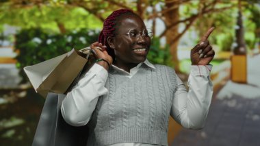 Parkta elinde alışveriş torbalarıyla gülümseyen, ağaçlara bakan, güneşli bir günün tadını çıkaran, beyaz gömlek ve gri kazak giyen, mutluluğu ifade eden, rahatlamış bir kadın..