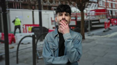 Genç bir adam açık hava inşaatında duruyor, kot ceket giyiyor, yakışıklı ve rahat poz veriyor..