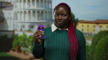 Güneşli bir günde elinde mor bir kurdele tutan Afrikalı Amerikalı kadın sidik kulesinin önünde duruyor, eşsiz bir miras ve ikonik İtalyan mimarisi kombinasyonunu vurguluyor..