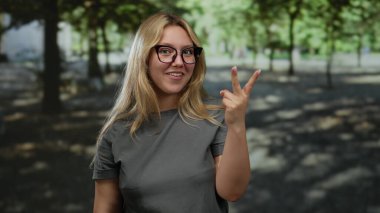 Parkta neşeli bir ifadeyle kutlama yapan, gözlük takan ve sıradan bir gömlek giyen, ağaçların altında bir anlık neşe yakalayan bir kadın..