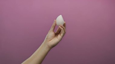 Beyaz adamın eli zarif bir şekilde canlı pembe bir arka plana karşı bir blender sünger tutuyor, tenini ve kozmetik ürünlerini izole bir ortamda vurguluyor..