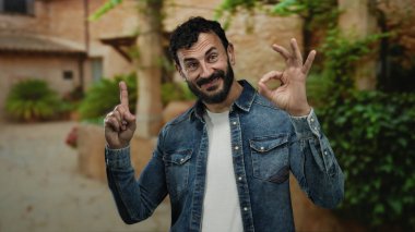 Sakallı İspanyol bir adam yeşil ağaçlarla çevrili güneşli bir parkta jest yapıyor. Kot gömlek giyiyor ve açık havada pozitifliğini gösteriyor..