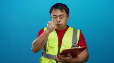 Emniyet yeleği giyen bir adam mavi arkaplan üzerinde pano tutarken tanımlı bir ifade gösteriyor.