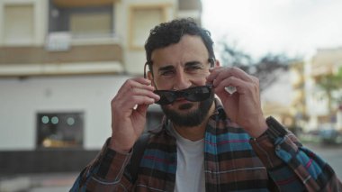 Sakallı, güneş gözlüğü takan İspanyol bir adam, açık bir gökyüzünün altında rahat bir açık hava havası yakalamış, gülümsüyor..