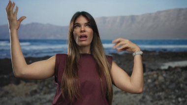 Genç İspanyol kadın, elleri kayalık bir plajda, arka planda dağlar ve okyanus ile duygusal hareketler yaparak, canlı bir açık hava sahnesini aktarıyor..