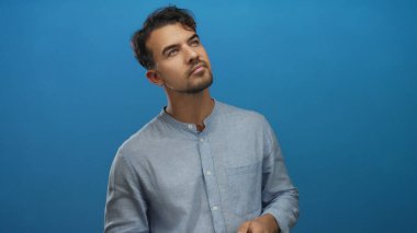 İzole edilmiş mavi arka plan karşısında kamerayla düşünen genç İspanyol bir adam, günlük gömlek giyiyor, yaratıcılığını ve düşüncelerini bir stüdyo ortamında aktarıyor..