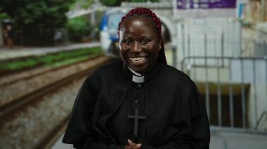 Tren durağında rahip kıyafetiyle gülen bir kadın. Arka planda bulanık bir tren var. Açık bir şehir ortamında neşe ve maneviyatı ifade ediyor..