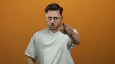 Sakallı genç adam, izole edilmiş turuncu arka planda kameraya kızgın bir şekilde el kol hareketi yapıyor. Günlük beyaz gömlek giyiyor..