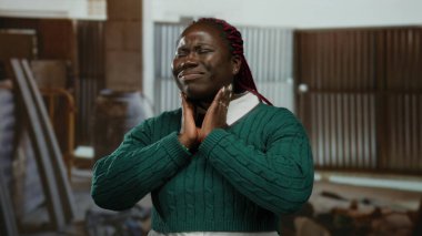 İnşaat alanında boynu ağrıyan, yeşil kazak giyen, iş ortamında endüstriyel aydınlatma altında rahatsızlığını ve yorgunluğunu ifade eden bir kadın..