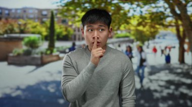 Genç adam, dudağını tutarak canlı bir şehir caddesinde sessizliği işaret ediyor. Arka planda bulanık insanlar ve ağaçlar var..