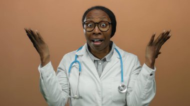 Kadın doktor şaşkınlığını ifade ediyor. Elleri kahverengi arka planda, gözlük ve steteskop takıyor.