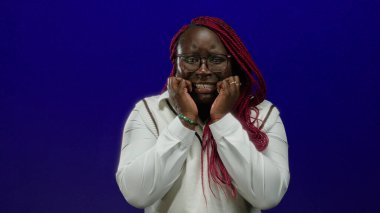 Kırmızı örgülü ve gözlüklü bir kadın mavi arka planda endişeli görünüyor. Endişelerini ya da endişelerini dile getiriyor..