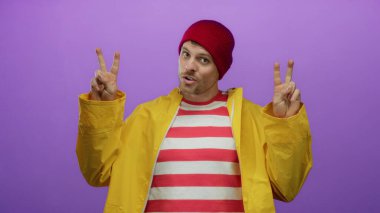 Sarı yağmurluk giyen, çizgili gömlekli genç adam canlı mor bir arka plana karşı jestler yapıyor..