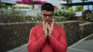 Kırmızı süveterli genç adam şehir sokaklarında dua ediyor. Arka planda canlı çiçekler var. Huzur ve yansıma hissi taşıyor..