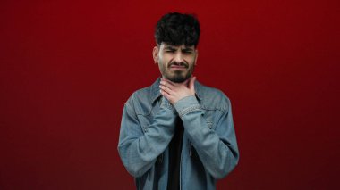 Kırmızı arka planda izole edilmiş genç adam üzgün görünüyor. Ellerini boğazına koyuyor. Kot ceket giyiyor.