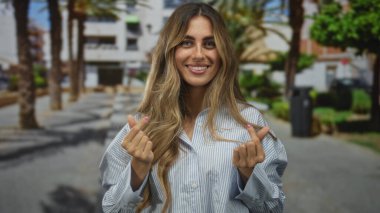 Uzun saçlı bir kadın, kaldırımda gülümserken iki eliyle el hareketi yapar..
