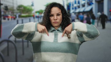 Yoğun caddede aşağı doğru hareket eden bir kadın şehir ortamında ciddi bir ifadeyle yön veya konumu belirterek kentsel ve gündelik tarzı harmanlıyor.