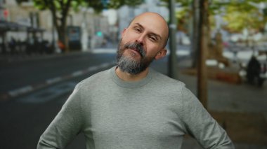 Şehir ortamında sakallı bir adam, gri süveteri ile ağaçlarla kaplı bir şehir sokağında güler yüzle, açık havada dostane bir hava tasvir ediyor..