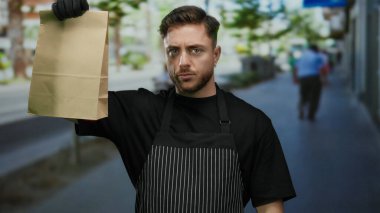 Eldiven giyen genç garson, şehir sokaklarında profesyonellik ve şehir canlılığını harmanlayan bir paket kâğıt çanta tutuyor..