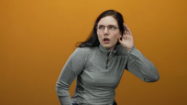 Gözlüklü bir kadın turuncu arka planda çeşitli yüz ifadeleri sergiliyor, gri bir kazak giyiyor, kafası karışmış görünüyor, dinliyor ve ilginç bir şekilde meşgul..