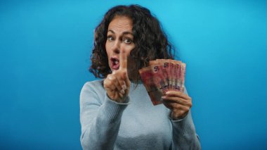 Öfkeyle el kol hareketi yapan bir kadın, Güney Afrika banknotlarını mavi duvara dayayıp para durumuyla ilgili hayal kırıklığını gösteriyor..