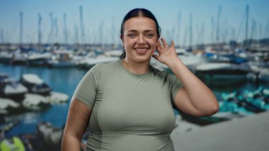 Arka planda tekneleri olan, deniz kıyısında gülümseyen genç ve cüsseli bir kadın, açık hava ve sakin atmosferin tadını çıkarıyor, pozitif ve kendinden emin bir tavır sergiliyor..