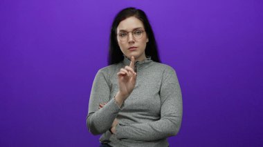Canlı mor arka plana karşı hareket eden, gözlük takan ve gri başlıklı kadın, onaylanmamış ifadeler ve odaklanmış ifadelerle sözlü olmayan iletişim kuruyor..