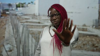 Gözlüklü ve kırmızı örgülü bir kadın arka planda bulanık bir şekilde inşaat alanında duruyor.