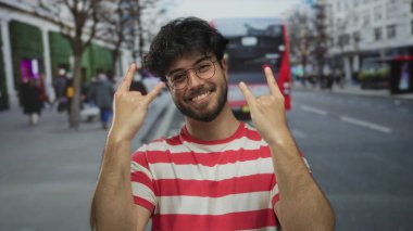Gözlüklü ve çizgili tişörtlü genç adam kalabalık şehir sokağında taştan jest yapıyor. Arka planda bulanık insanlar ve otobüsler, açık havada gençlik enerjisi ve mutluluk sergiliyor..