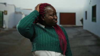 Şehir merkezinde yeşil kazak giyen pembe örgülü bir kadın sıradan bir ortamda sakin bir yüz ifadesi sergiliyor..