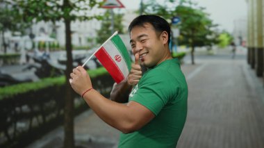 Başparmaklarıyla gülümseyen genç adam İran bayrağını şehir caddesinde tutuyor.