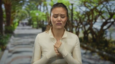 Parktaki İspanyol kadın duygusal görünüyor. Arka planda yeşil yapraklar var. Açık renk ceket giyiyor. Üzüntüsünü dile getiriyor..