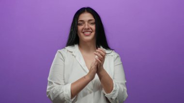 İzole edilmiş mor arka planda, beyaz gömlek giyen, kendine güveni ve pozitifliğiyle mutlu bir şekilde alkışlayan kadın..