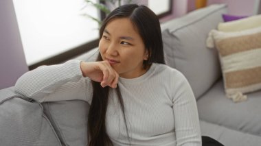 Bir dairedeki rahat bir koltukta dinlenen bir kadın, modern bir oturma odasında sakin ve düşünceli titreşimler yayıyor, hafif ışıklandırma ve gündelik dekorla..