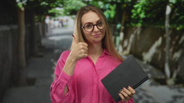 Elinde kitap tutan genç bir kadın şehir sokağında yeşil ağaçlarla çevrili gözlüklü ve pembe tişörtlü bir şekilde olumlu ve kendine güvenini gösteren bir aferin veriyor..