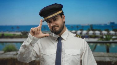 Pilot üniformalı İspanyol sakallı adam arka planda sakin mavi deniz ve palmiye ağaçlarıyla deniz kenarında ezik jestler yapıyor..