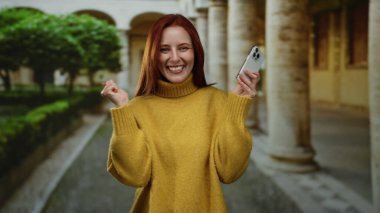 Üniversite avlusunda elinde akıllı telefonla gülümseyen bir kadın, tarihi mimari arka planda günlük sarı süveter giymiş kızıl saçlı güzelle neşe ve modern iletişim sergiliyor..