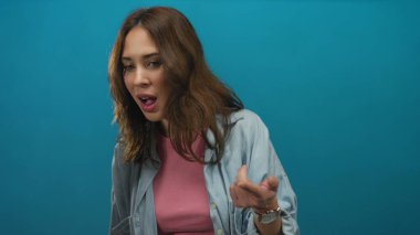 Latin kökenli genç bir kadın, canlı mavi bir arkaplanda izole edilmiş, günlük giysiler giymiş ve görünmeyen bir şeyi sorguluyormuş gibi el kol hareketi yapıyor..