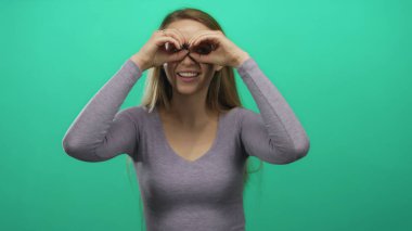 Uzun saçlı genç kadın, yeşil arka plan duvarına karşı elleri dürbünlü dürbünü taklit ediyor.