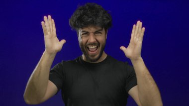 Sakallı genç İspanyol bir adam mavi arka planda hayal kırıklığı yaşıyor, elleri yoğun bir duygu ve enerji gösterisi içinde..