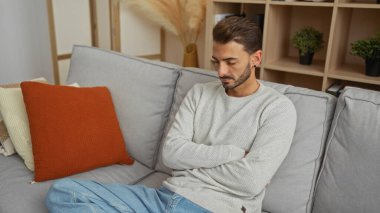 Modern bir apartman dairesinde kolları çaprazlanmış, koltukta oturan genç İspanyol bir adam. İçinde bitkiler ve hoş bir dekor olan, iç gözlem havasını iletiyor.