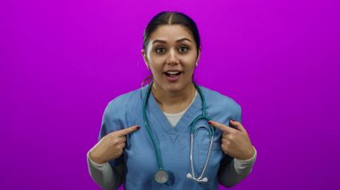 Mavi önlüklü, steteskoplu İspanyol hemşire, kendini izole edilmiş pembe duvar arka planına işaret ederek şaşırmış görünüyor..