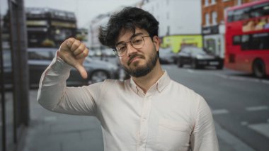 Sakallı genç adam dışarıda, kalabalık bir sokakta arka planda arabaları ve otobüsleri olan şehir ortamında gözlüklü ve beyaz gömlekli bir jest yapıyor..