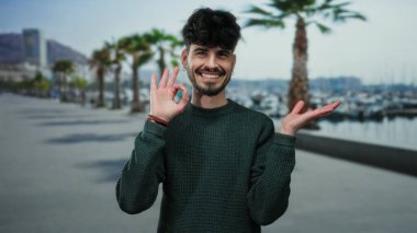 Gülümseyen süveter içindeki genç adam, açık gökyüzünün altında palmiye ağaçları ve marina arkaplanı olan deniz kenarındaki bir geçidi işaret ederek modern gündelik yaşam tarzını resmediyor..