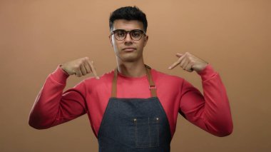 Gözlüklü, kırmızı tişörtlü ve kot önlüklü genç adam kahverengi arka planda kendine işaret ediyor..