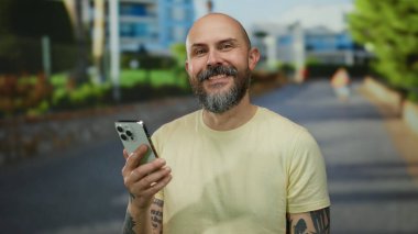 Sarı tişörtlü kel adam şehir sokağında elinde dövmeli akıllı bir telefonla duruyor. Açık havada kendine güvenen ve ulaşılabilir görünüyor..