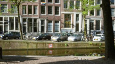 Amsterdam 'ın ikonik kanallarının bulanık görüntüsü. Parlak gün ışığında odakları kesilmiş arabalar, bisikletler ve geleneksel Hollanda mimarisi..