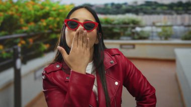 Şık kırmızı ceketli ve güneş gözlüklü genç bir kadın açık havada canlı yeşil arka planlı bir öpücük yolluyor..